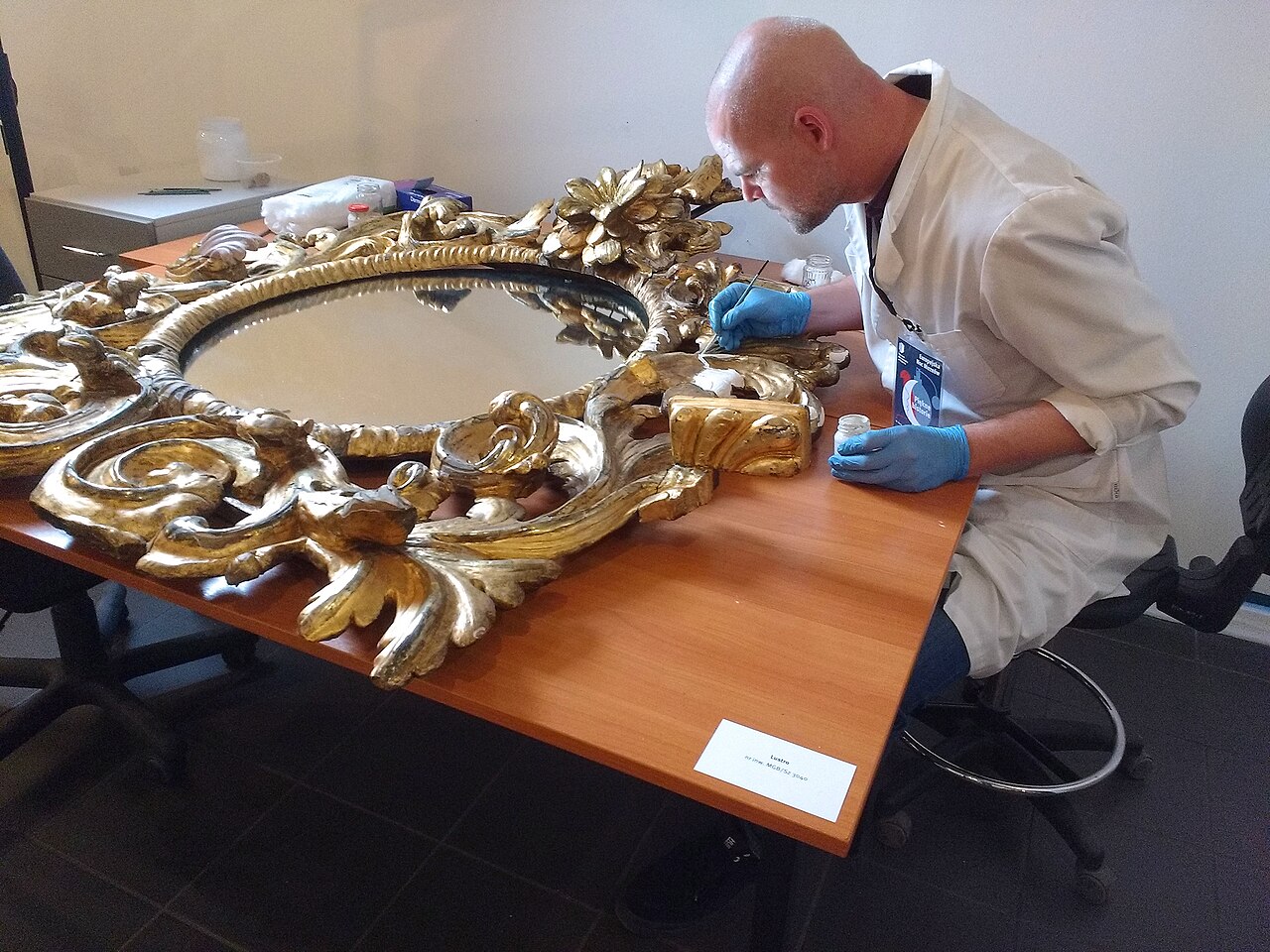 Реставратор в халате восстанавливает золочёную резную раму старинного зеркала на столе